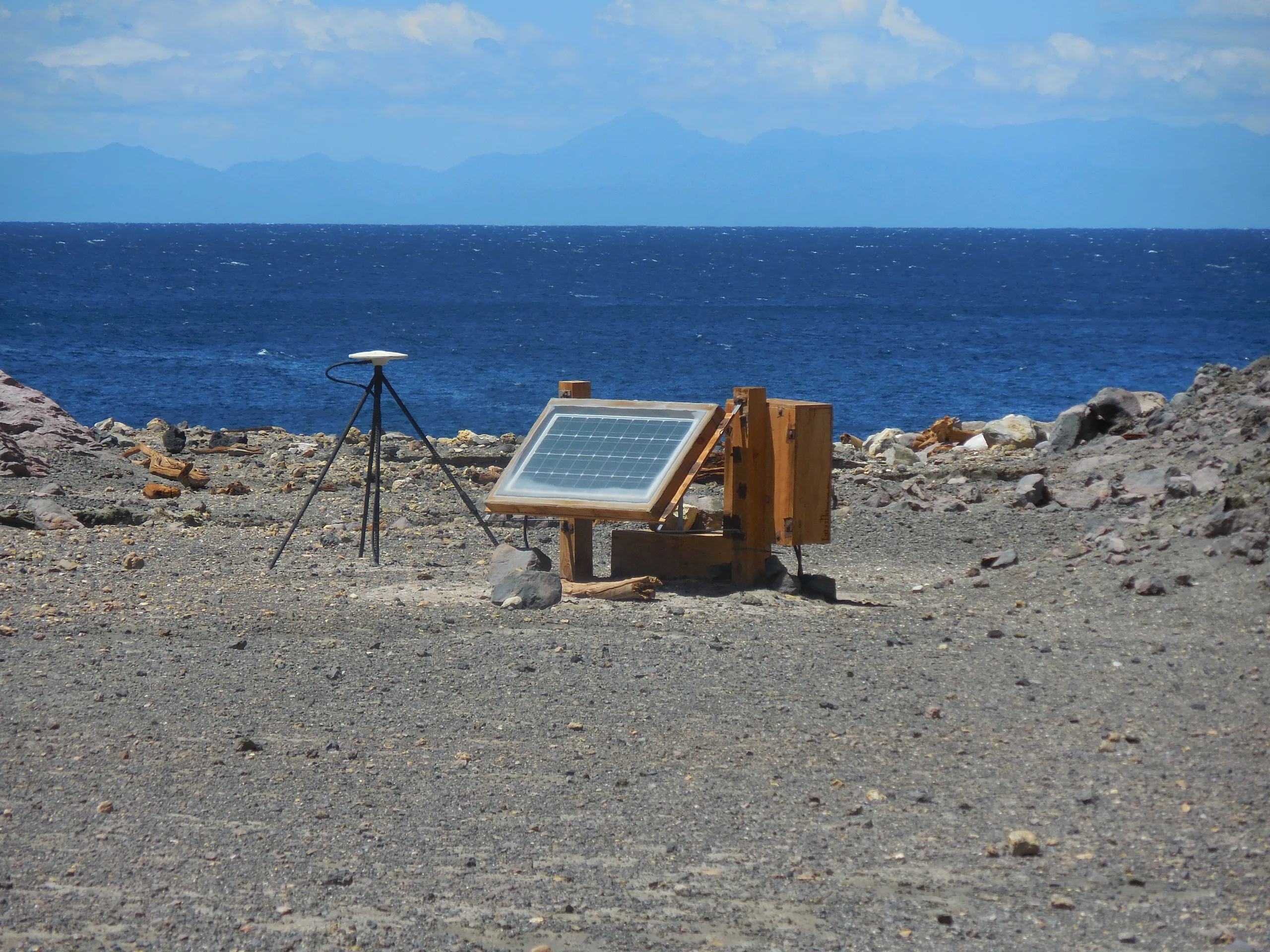 A solar-powered volcano monitoring station with a GNSS antenna on the bare volcanic ground of White Island, the ocean behind it